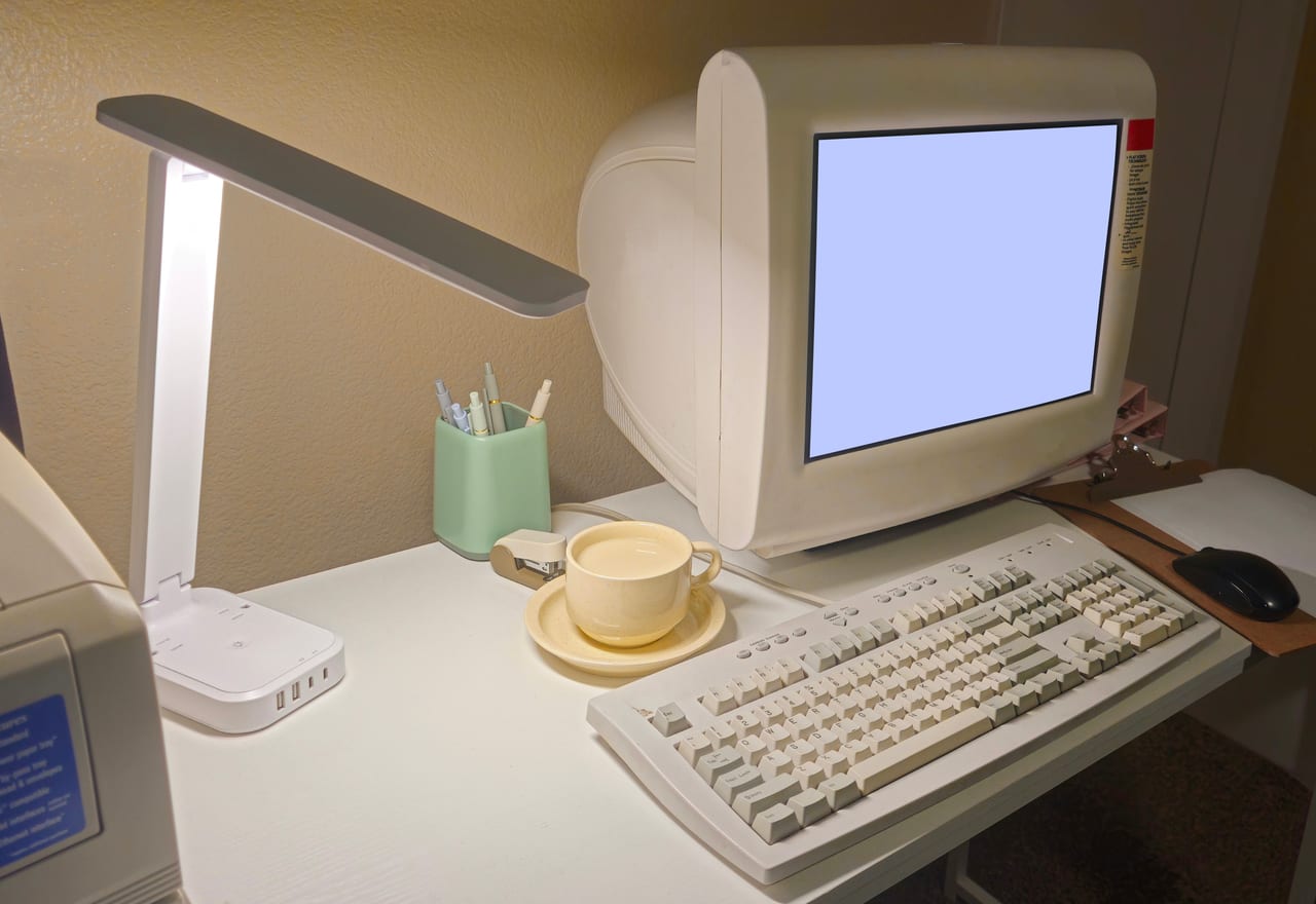 Alter Röhrenmonitor mit beiger Tastatur auf einem Schreibtisch, daneben Kaffeetasse und Schreibtischlampe