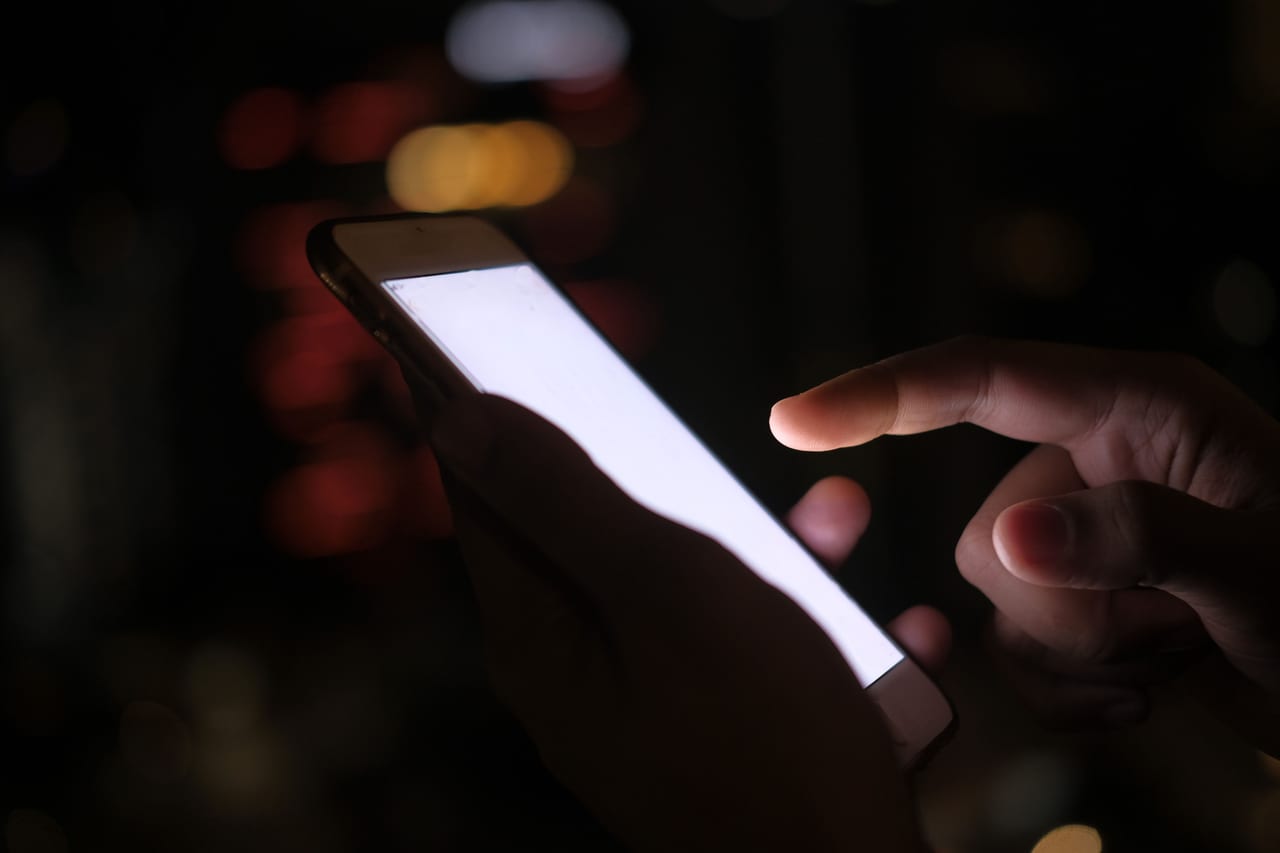Hand tippt im Dunkeln auf ein hell leuchtendes Smartphone, im Hintergrund unscharfe Lichter