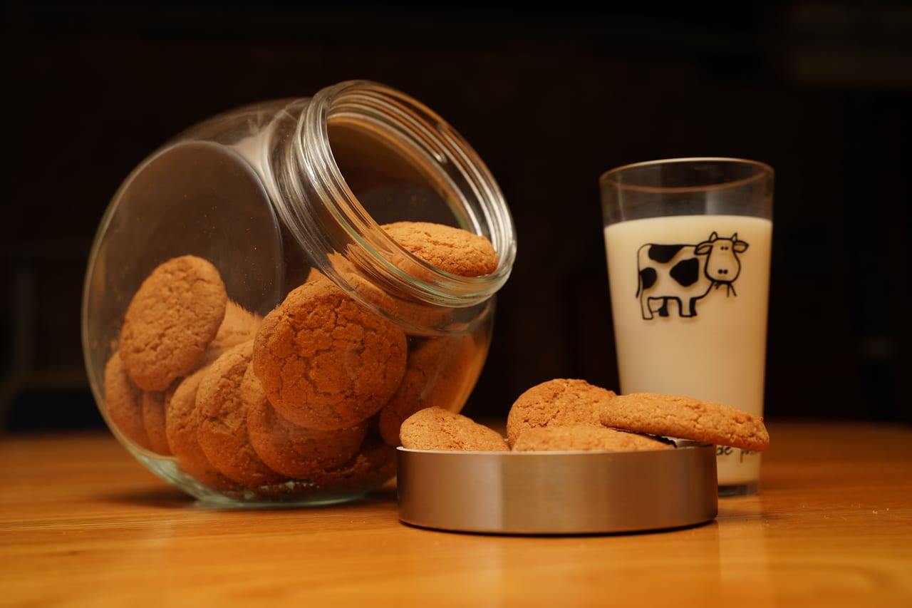 Umgekipptes Glas mit Haferkeksen neben dem Deckel voller Kekse und einem Glas Milch auf einem Holztisch