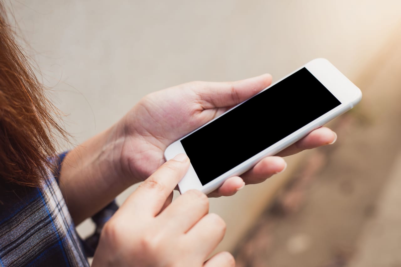Zwei Hände halten ein weißes Smartphone mit dunklem Bildschirm, ein Finger tippt auf das Display