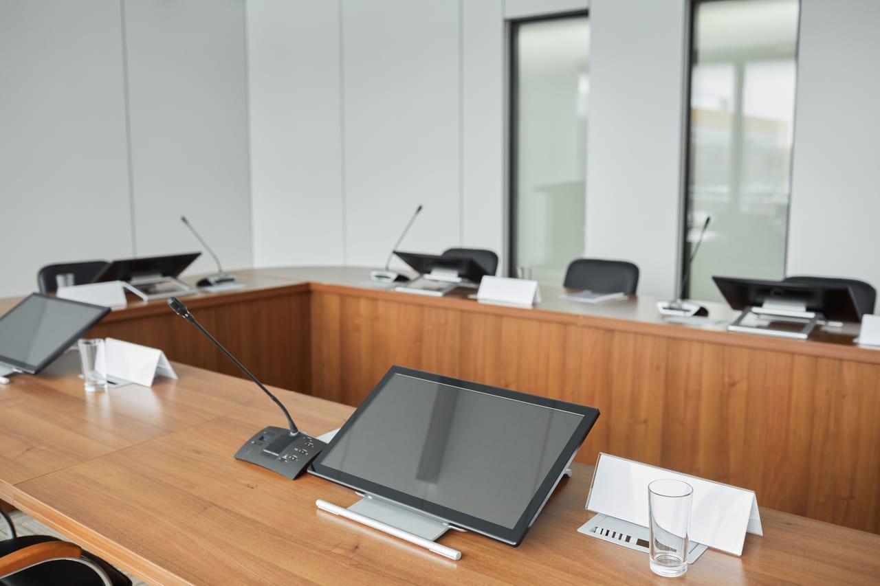 Konferenzraum mit langem Tisch, Mikrofonen an jedem Platz und Bildschirmen an der Stirnseite