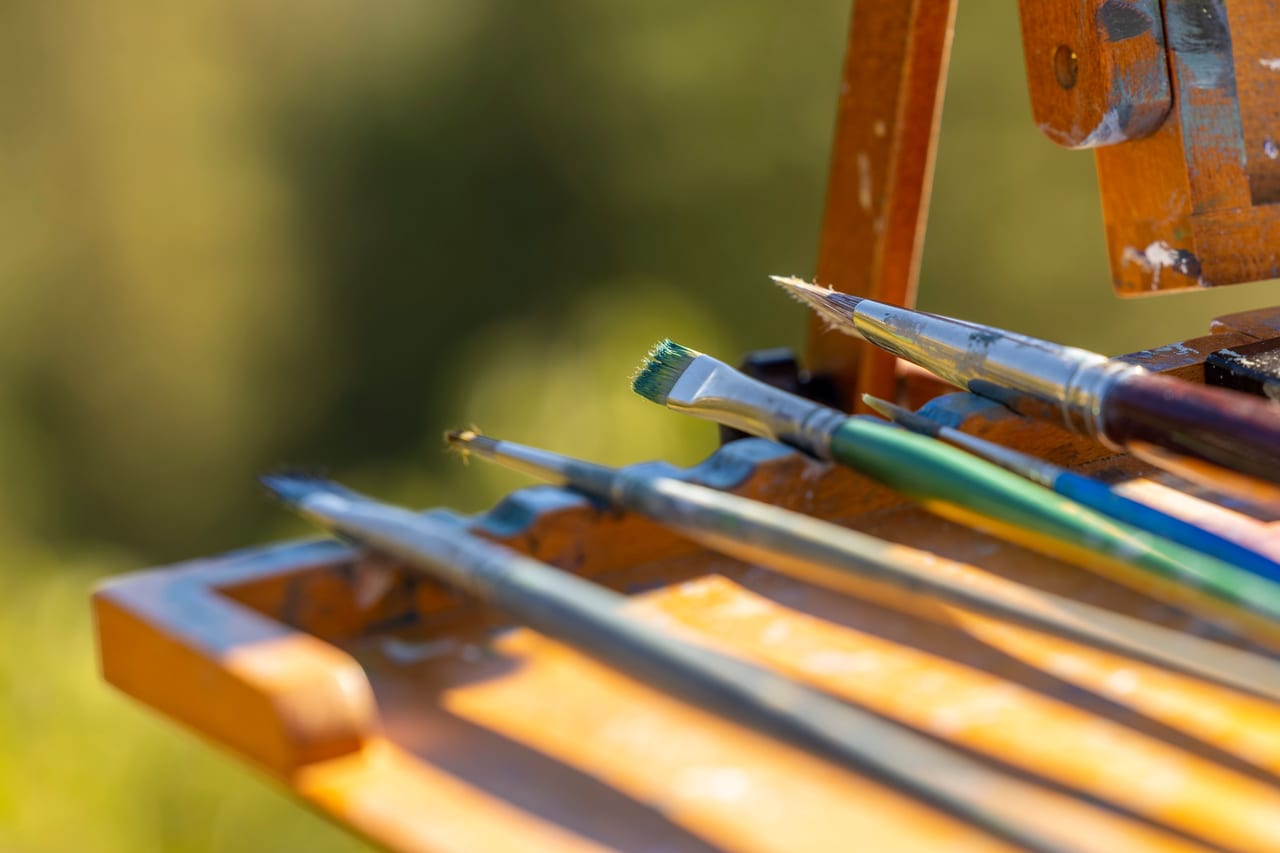 Benutzte Pinsel mit Farbresten liegen in der Ablage einer hölzernen Staffelei im Sonnenlicht