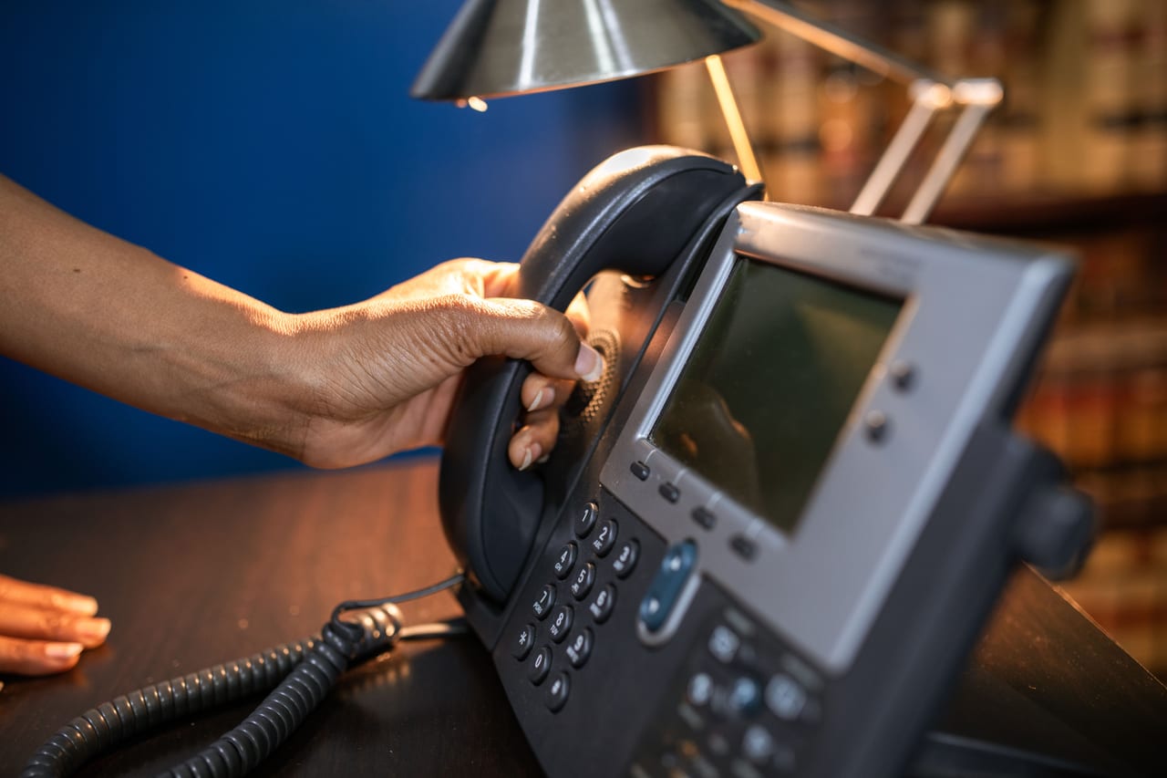 Hand nimmt den Hörer eines Bürotelefons mit Display ab, Schreibtischlampe im Hintergrund