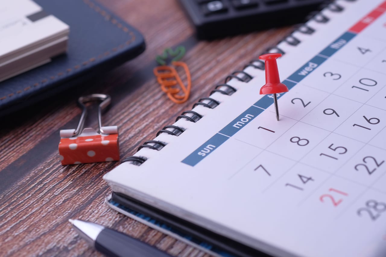 Rote Pinnnadel steckt in einem Ringkalender auf einem Holzschreibtisch, daneben Büroklammer und Stift