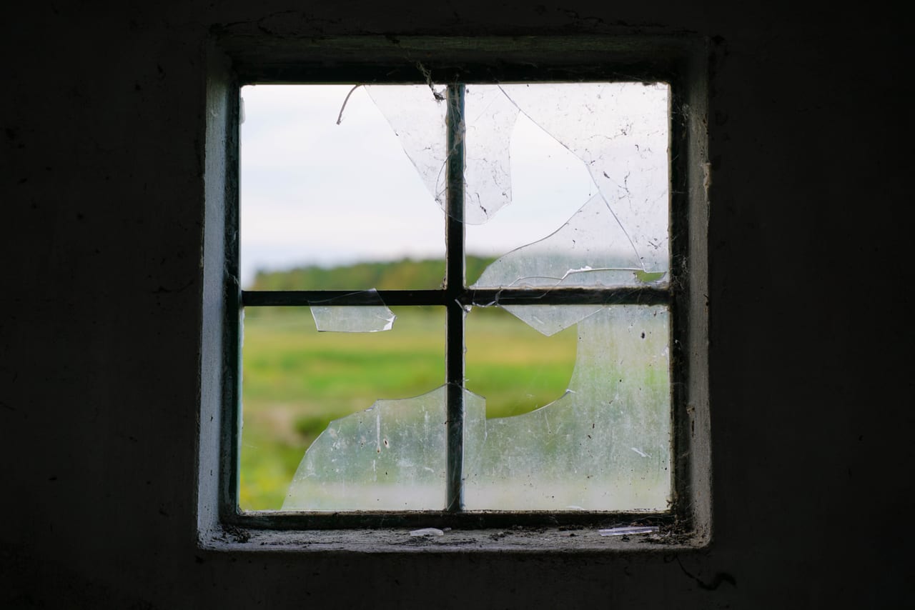 Kleines Sprossenfenster mit zerbrochener Scheibe in einer dunklen Wand, dahinter eine grüne Wiese
