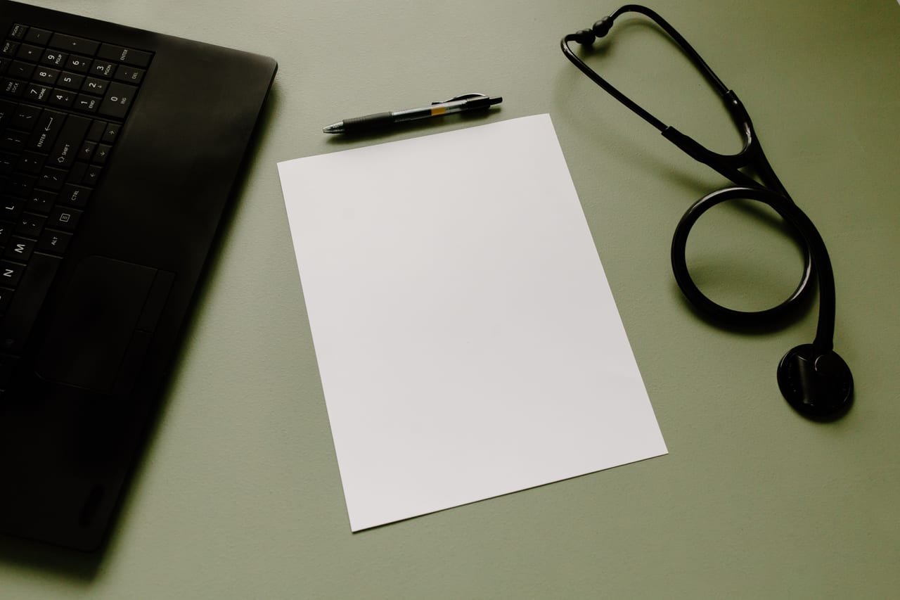 Aufgeklappter Laptop, leeres Blatt Papier, Kugelschreiber und Stethoskop auf einem olivgrünen Schreibtisch