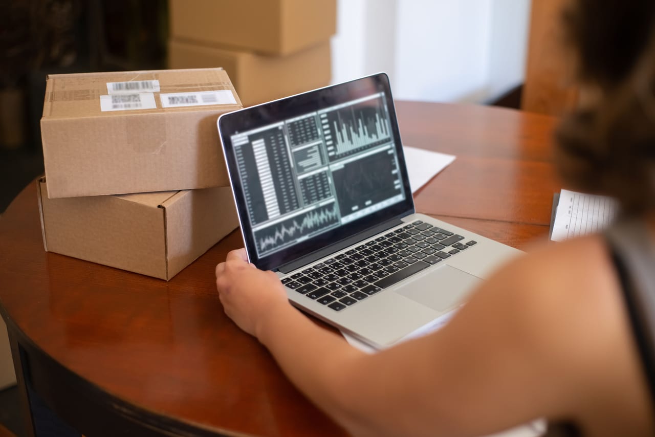 Versandkartons mit Barcode-Etiketten neben einem Laptop, auf dem Verkaufsstatistiken angezeigt werden