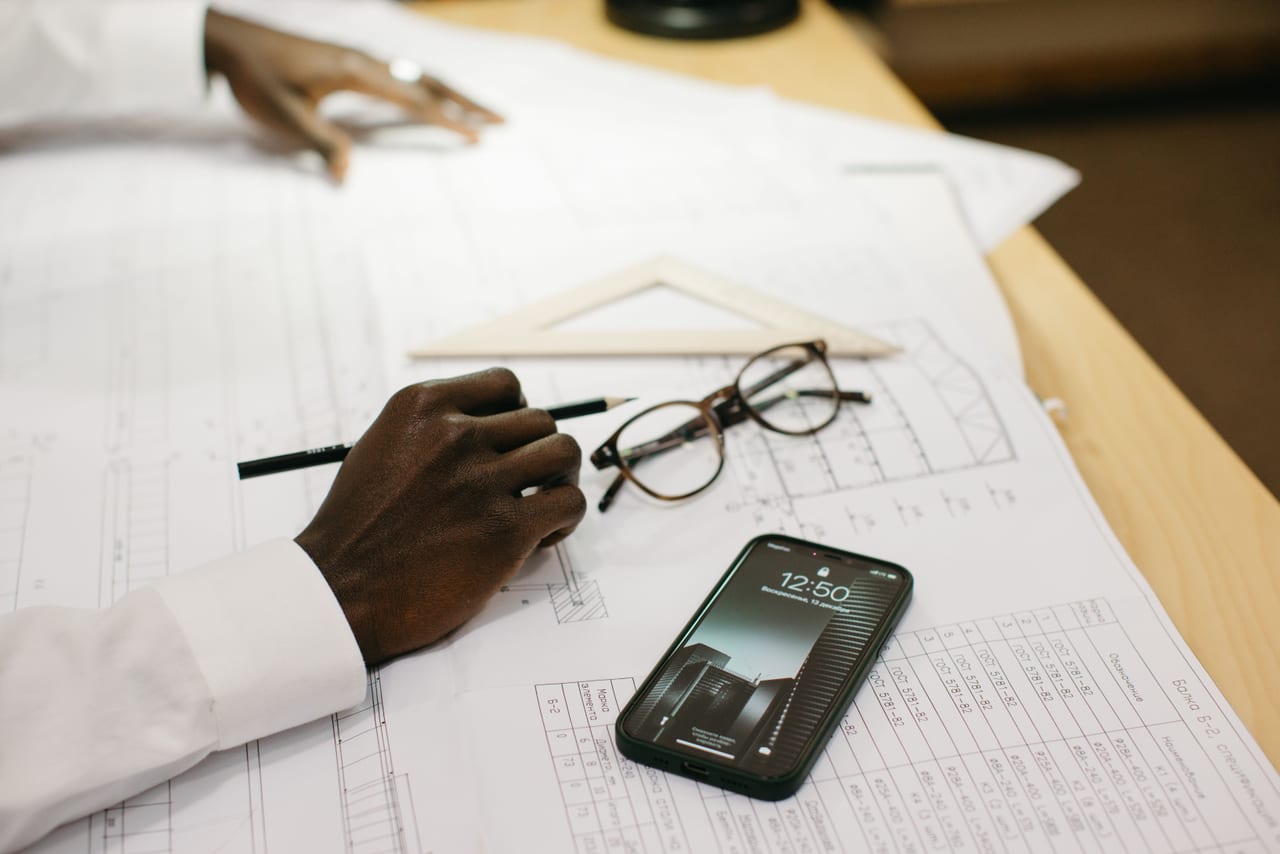Hände mit Bleistift über einem Bauplan, daneben Geodreieck, Brille und Smartphone