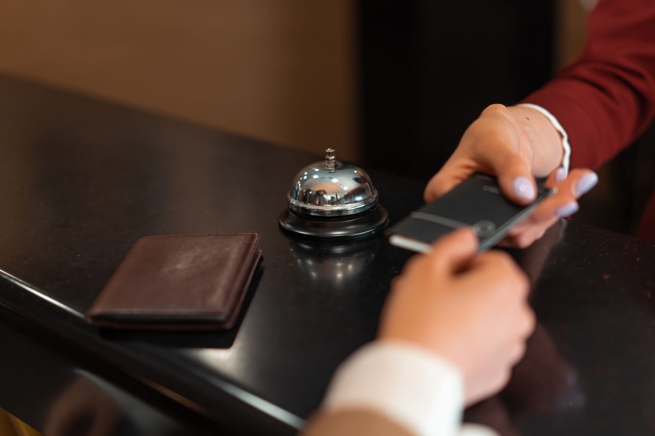 Dunkle Hotelrezeption mit Tischklingel, davor Hände über einem Smartphone