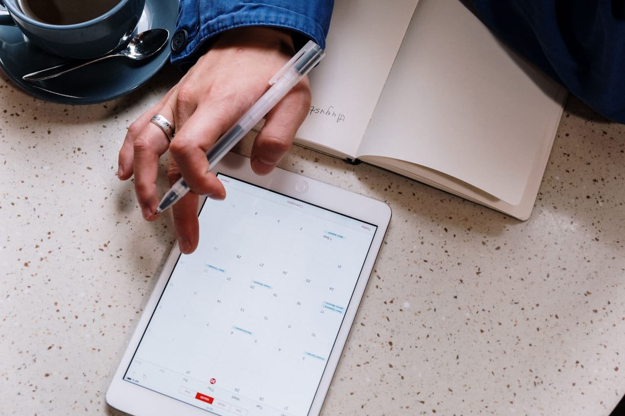 Hand mit Stift über einem Tablet, das eine Kalender-Monatsansicht mit eingetragenen Terminen zeigt, daneben ein Notizbuch