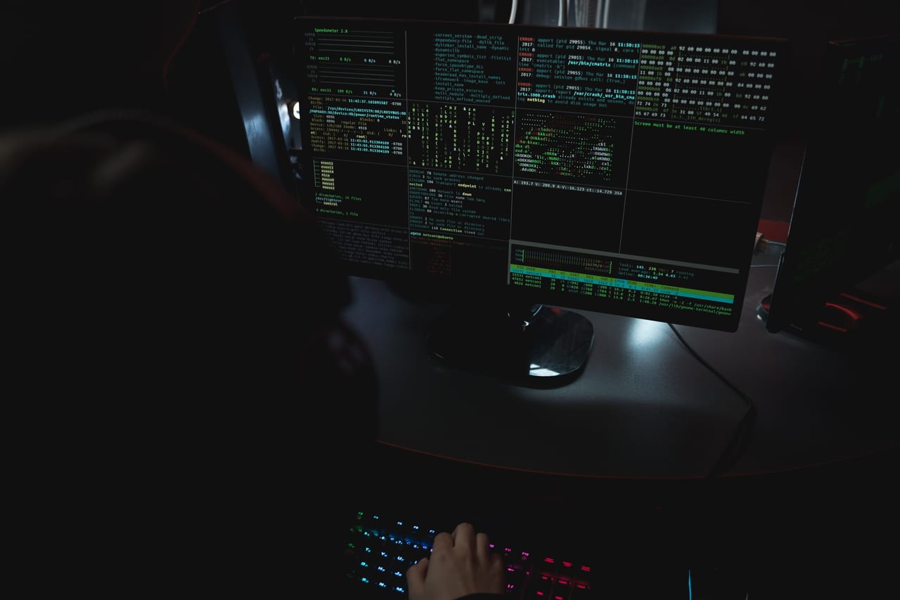 Zwei Bildschirme mit Quelltext und Systemanzeigen in einem abgedunkelten Raum, davor eine Tastatur