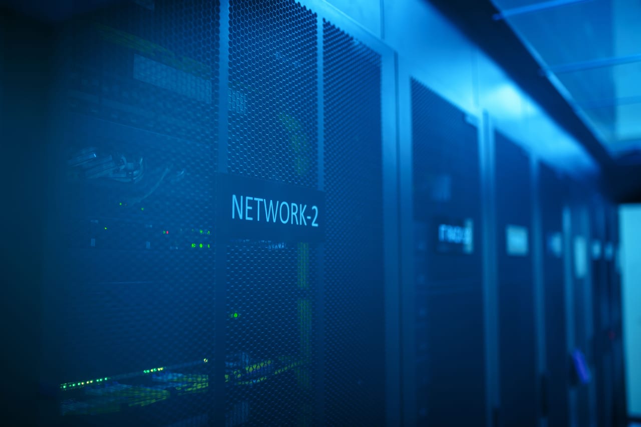 Reihe geschlossener Serverschränke in einem blau beleuchteten Rechenzentrum, an einem Schrank das Schild „NETWORK-2“