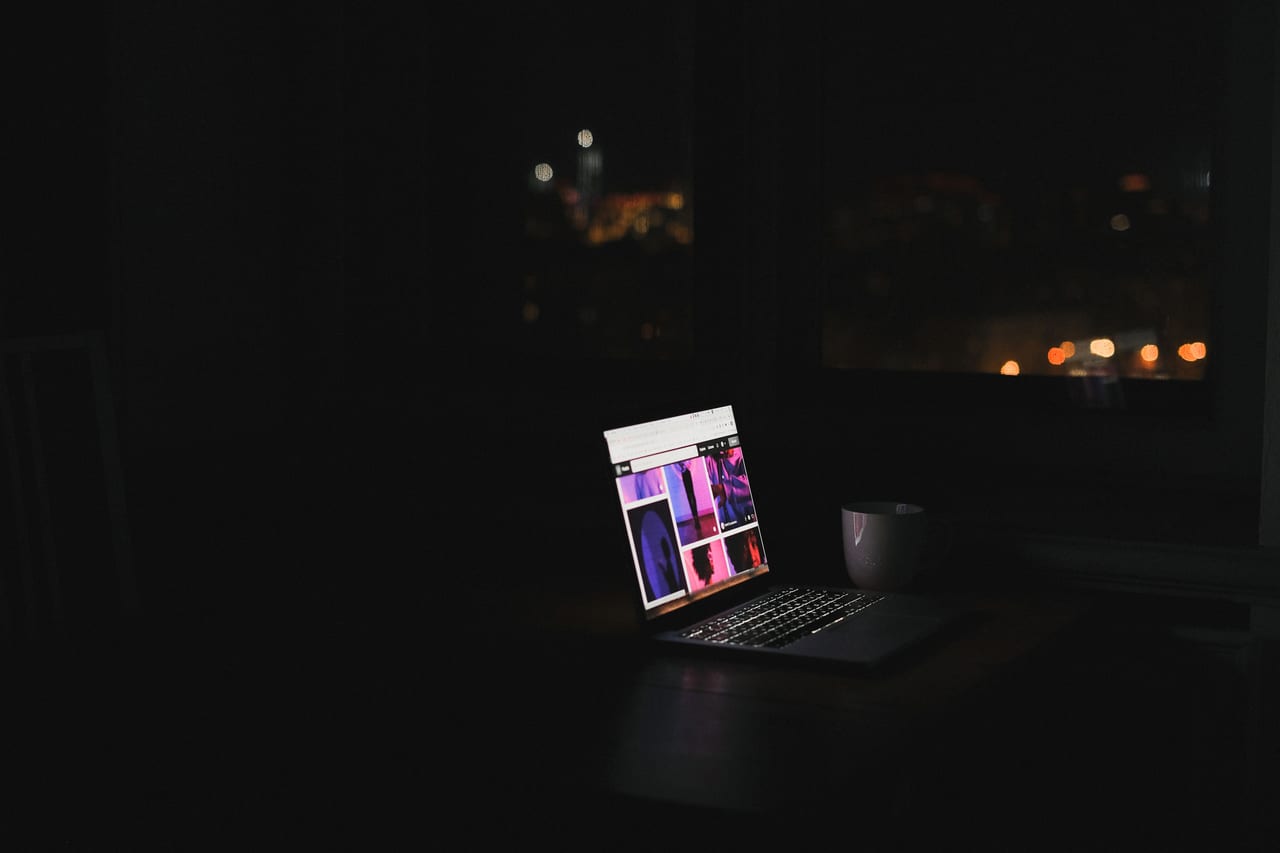 Aufgeklapptes Notebook leuchtet in einem dunklen Raum, dahinter die Lichter einer Stadt im Fenster