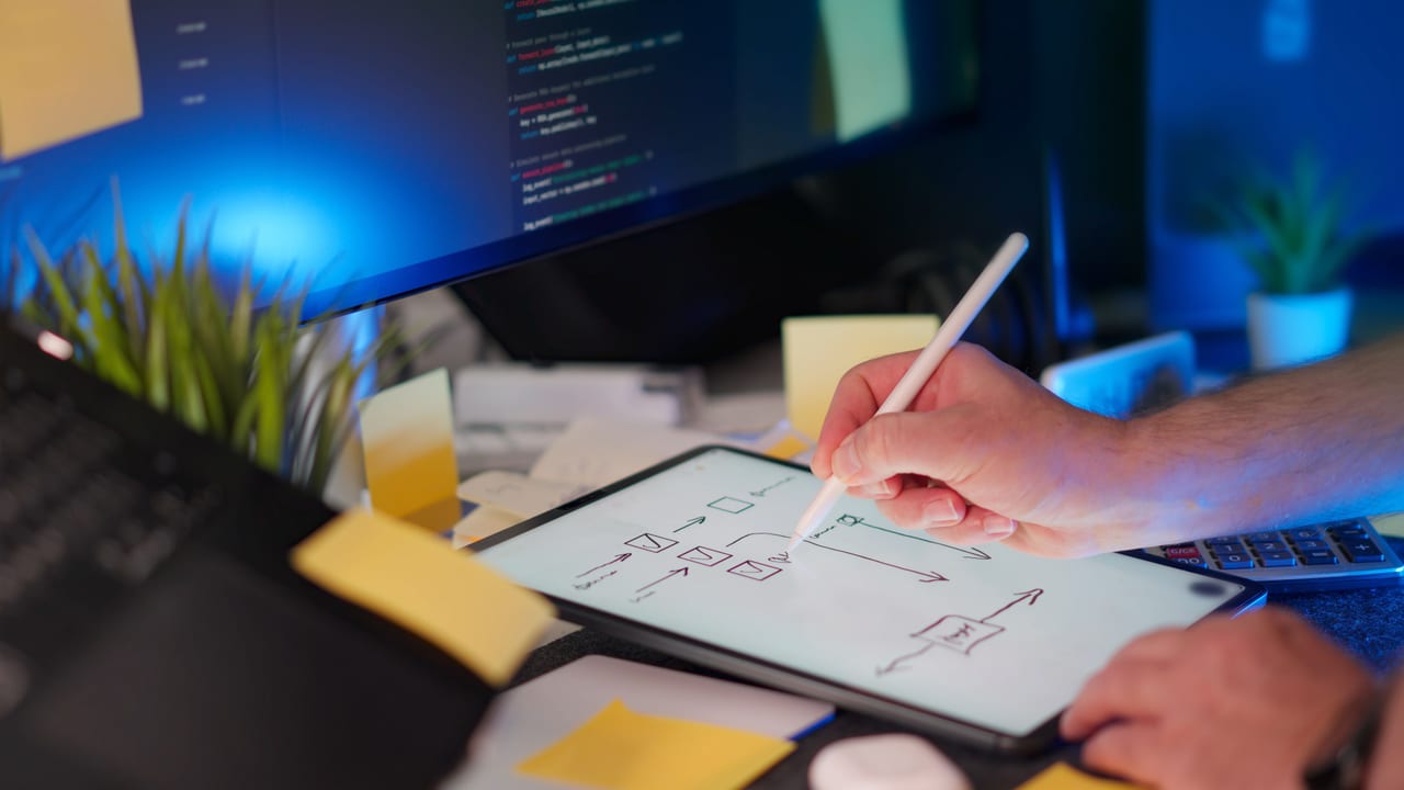 Hand zeichnet mit Stift ein Blockdiagramm mit Pfeilen auf einem Tablet, im Hintergrund Quellcode auf einem Monitor