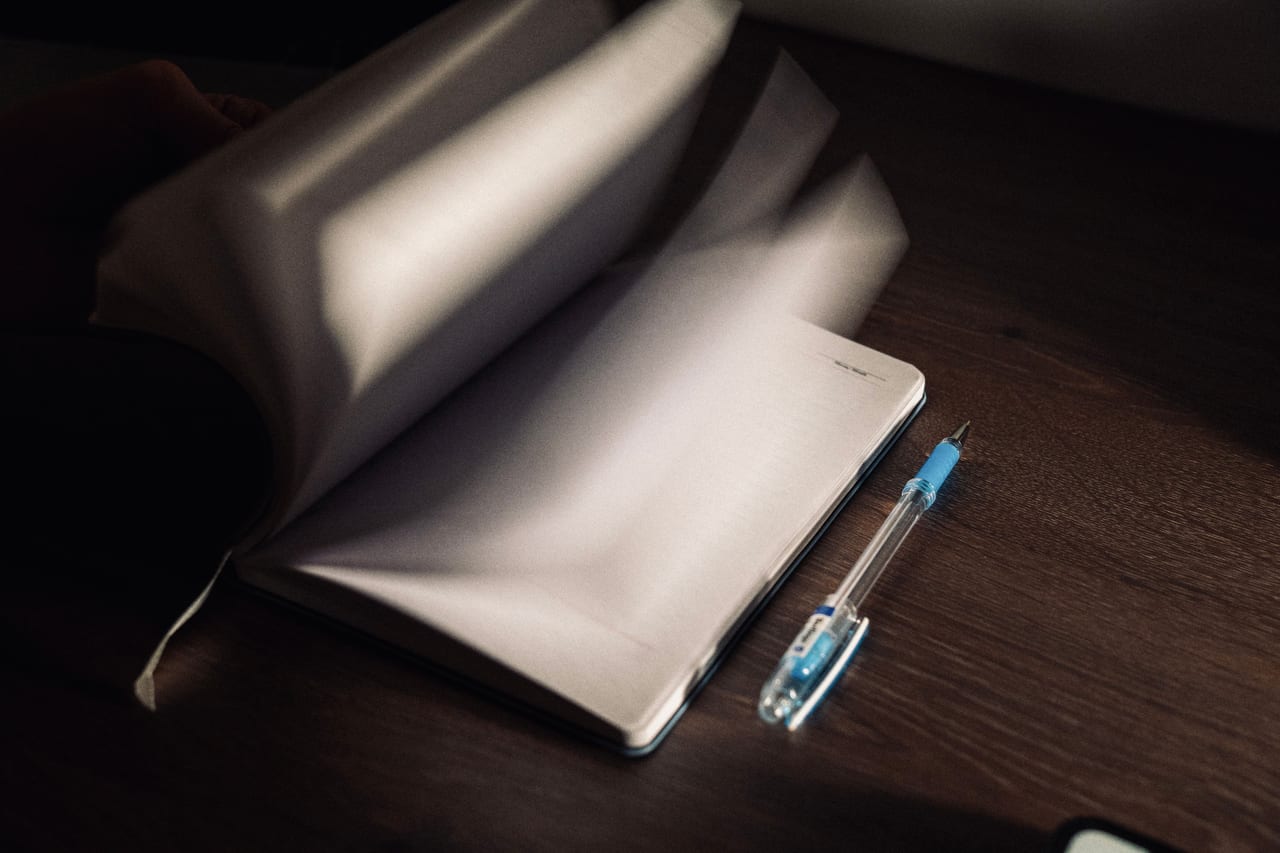 Aufgeschlagenes Notizbuch mit hellen Seiten und ein blauer Kugelschreiber auf einem dunklen Holztisch