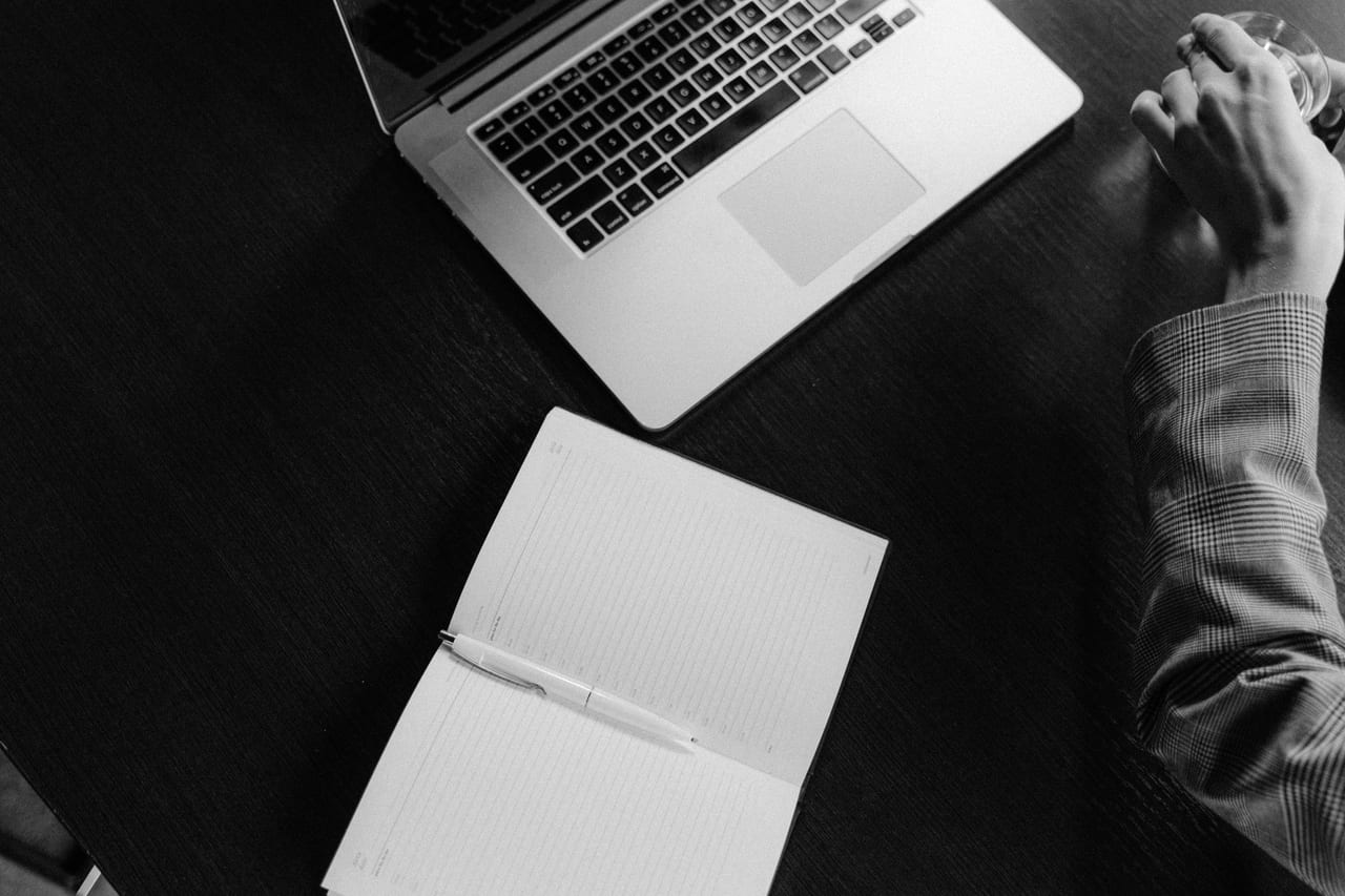 Schwarzweiß: aufgeklapptes Notebook und ein Notizbuch mit Stift auf einer dunklen Tischplatte