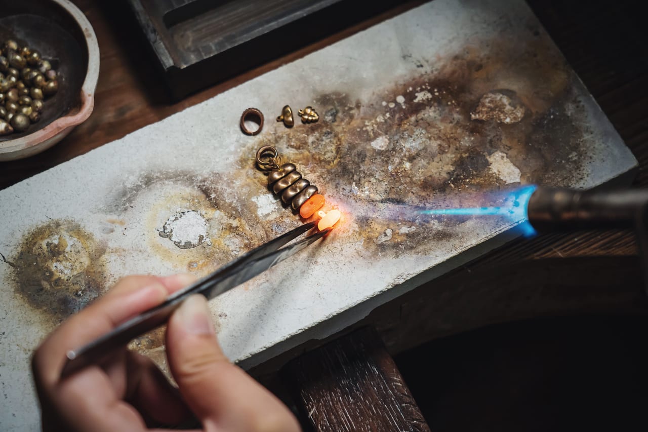 Hand hält mit einer Pinzette ein Schmuckstück in die Flamme eines Lötbrenners auf einer verrußten Lötplatte